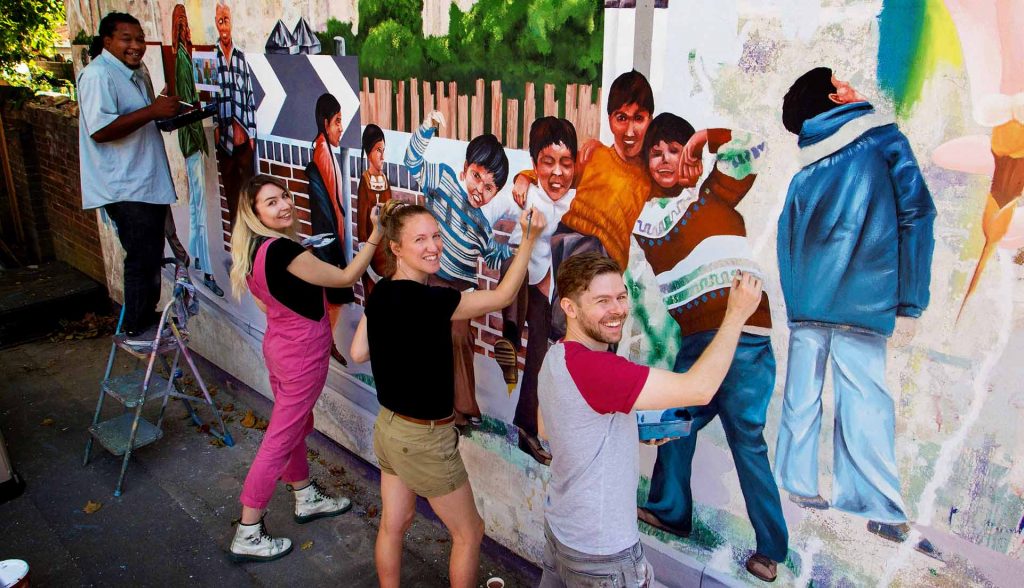 Three artists stood in a line, painting a colourful mural on a wall. The artists are looking up to the camera and smiling, while posing holding their brushes on the wall.