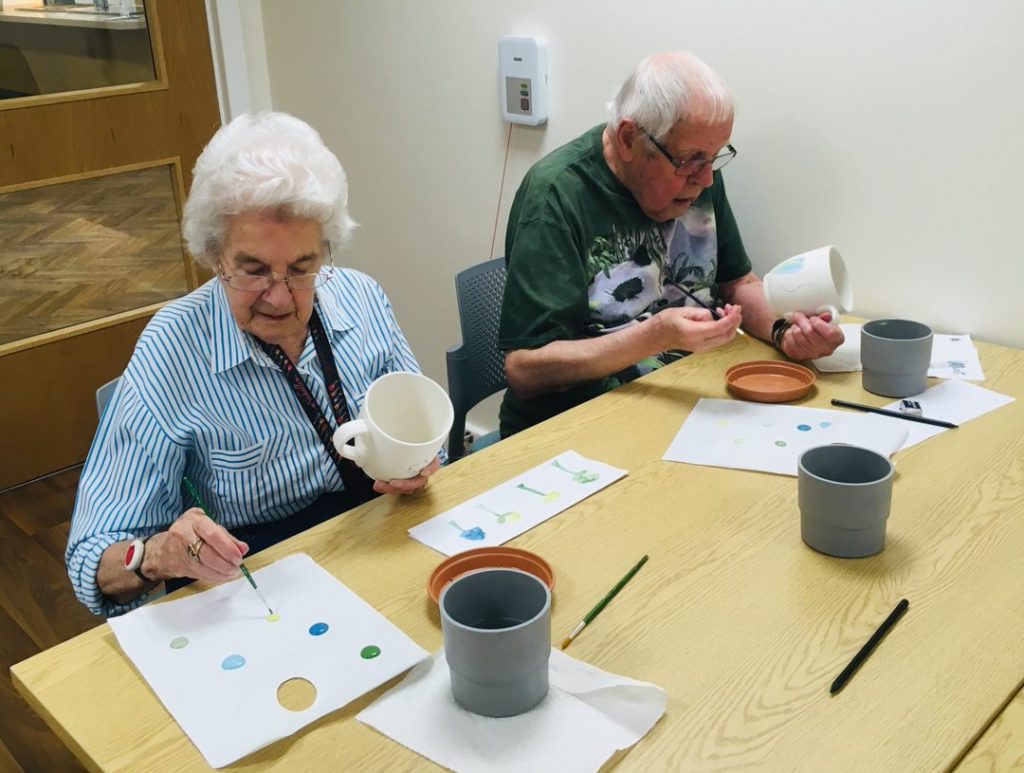 a woman and a man sit at a table, each has a paint brush and is painting shapes onto a blank cup. One the table there are water pots and spare brushes. Both look engrossed in what they are doing