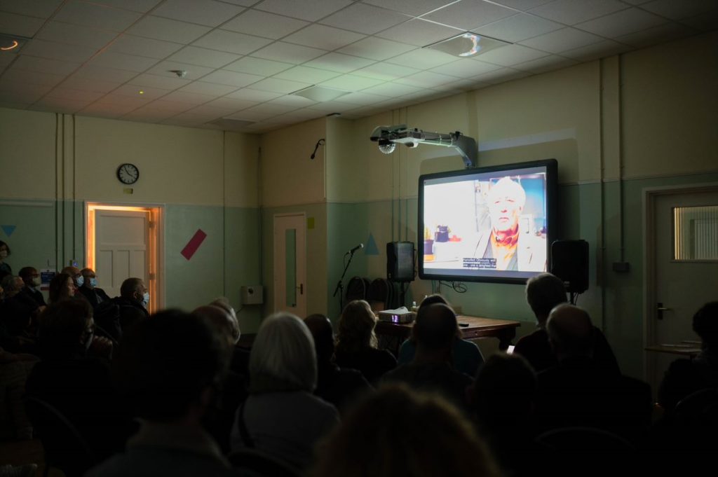 A dark photo of a crowd of people watching a documentary on a projector screen.