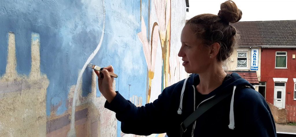 Mural artist Amanda painting a top section of the mural. She is looking at the mural and holding a paintbrush to the wall.