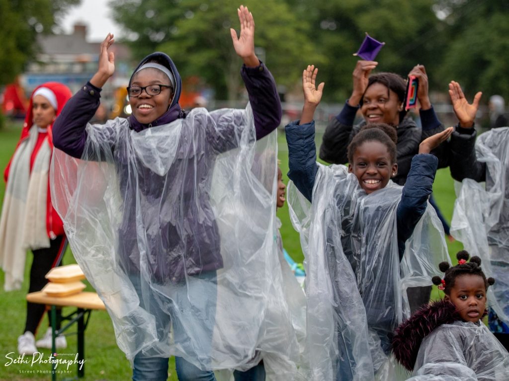 A group of Millfield festival attendees cheering. They are all wearing plastic rain ponchos on top of coats. All have their arms raised and cheerful expressions.