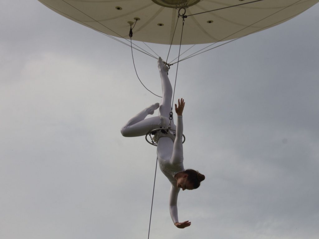 A photo of an aerial performance. A ballerina hangs upside down, attached to a hot air balloon.