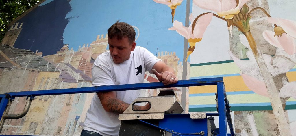 Mural artist Nathan on a cherrypicker, looking down at the ground. A section of a half-painted mural is behind him.