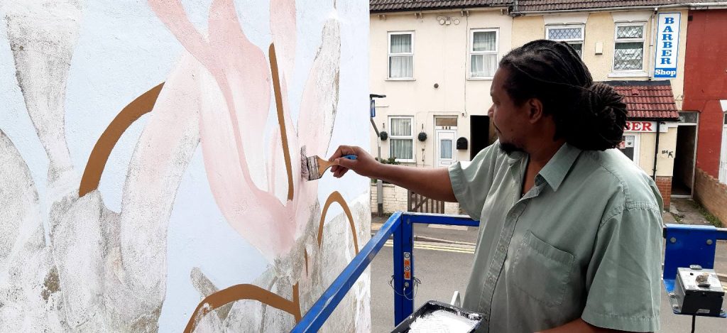 Mural artist Tony painting a section of the mural. He is stood on a cherrypicker and holding a paintbrush to the wall.