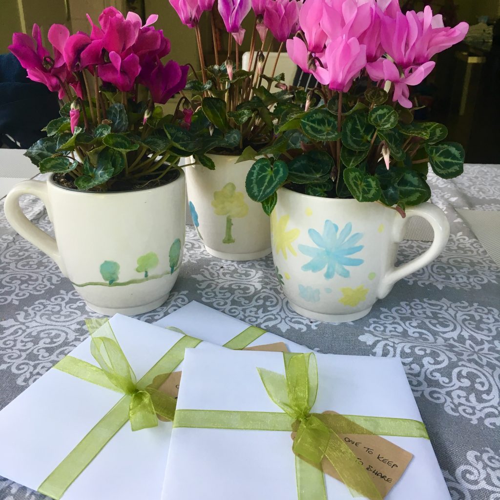 three white cups with colourful painted designs on them contain plants. The plants have green foliage and purple flowers. In front of the are three flat square parcels, each with gold ribbons on them and a brown gift tag