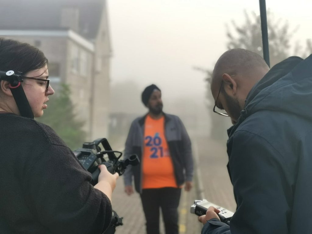 A behind the scenes photo of Deljit recording his memory tours. Deljit is wearing a neon orange top, and is in the centre of the frame but flurry and shrouded in fog. Emily and Os are stood with their backs to the camera, looking at their camera and sound equipment.