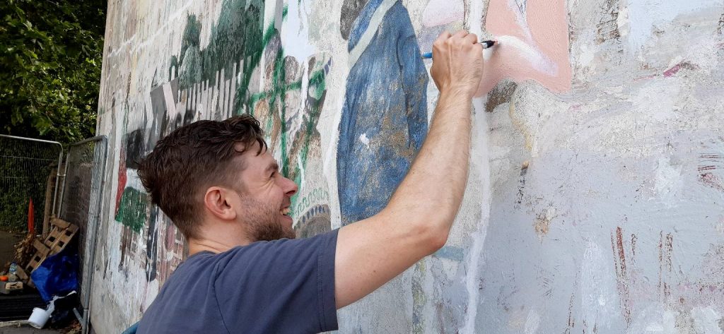 A photo of one of the mural artists, Jason, beginning to repaint the mural. Jason is looking at the wall and holding a paintbrush at eye-level on the wall.
