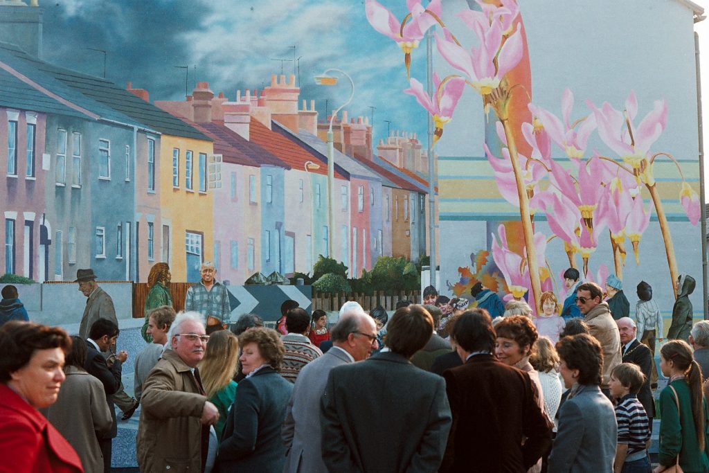A photograph taken in the 1980s of a crowd of people stood in front of a colourful mural.