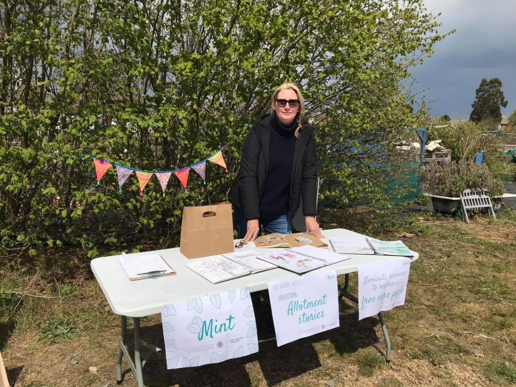 a woman in a black jacket leans on a table, facing the camera. Behind her is a trail of bunting. The table has posters hanging off the front with the words Mint on them. She is on an allotment site