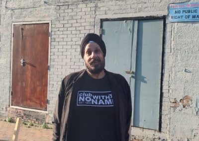 A photo of Deljit Singh stood in front of the former The Park music venue. Del is wearing a black turban, a black zipped jacket (worn open), and a black t-shirt which reads: the club with no name. Del is looking into the camera and smiling