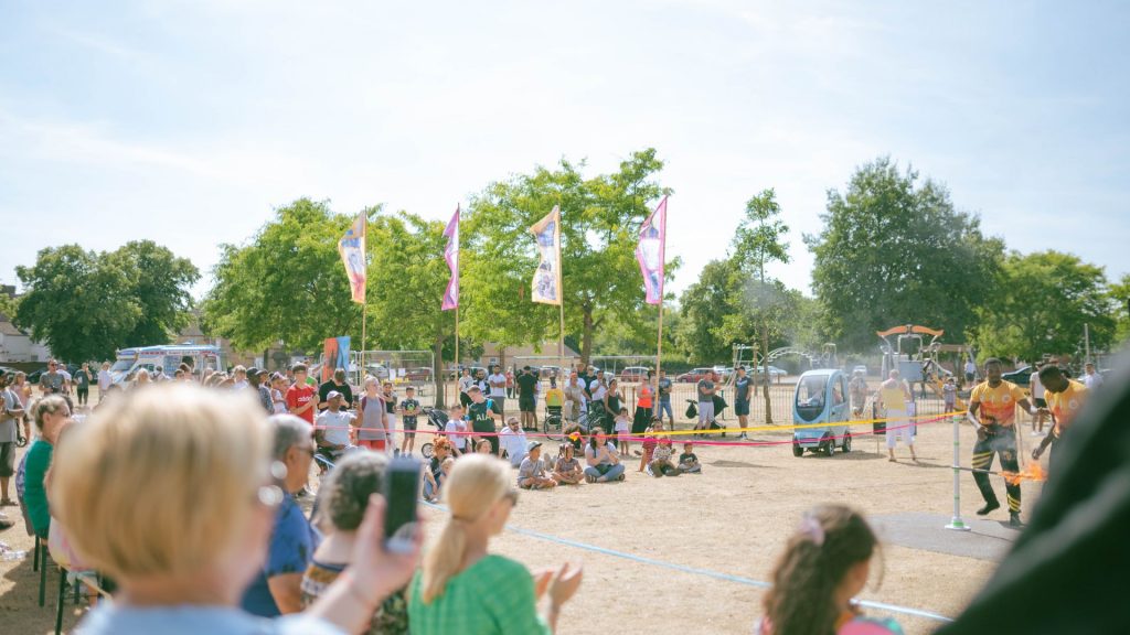 A photo of a crowd watching the Black Eagles acrobatics performance. The festival is outside in a field. Four large flags are in the background, in front of some trees.