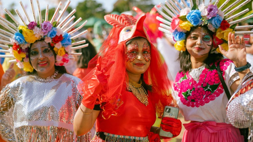 A photo of three people at Millfield Carnival. They are wearing brightly coloured costumes. The person in the centre is dressed as a red bird and the people either side have flowery headdresses and sparkly outfits. They are all looking down the lens and smiling.