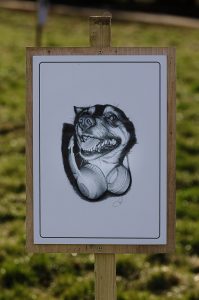 A realistic drawing of a staffie-mix, on display on a wooden sign thats fixed into the grass by a wooden post
