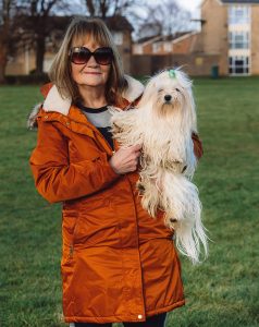 A woman in an orange coat and sunglasses holding her long-haired maltese dog. Stood on the grass. The event was WestWoof dog walk and social in Westwood, Peterborough.