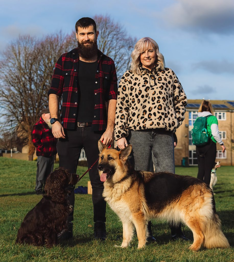 A couple pose with their two dogs, a spaniel and a german shepherd. Stood on the grass. The event was WestWoof dog walk and social in Westwood, Peterborough.