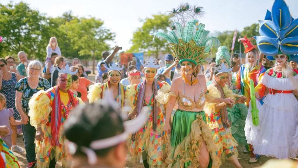 The Carnival in full swing at Millfield Festival 2023. crowds of people around the Carnival people in full colourful costume including hats and head gear.