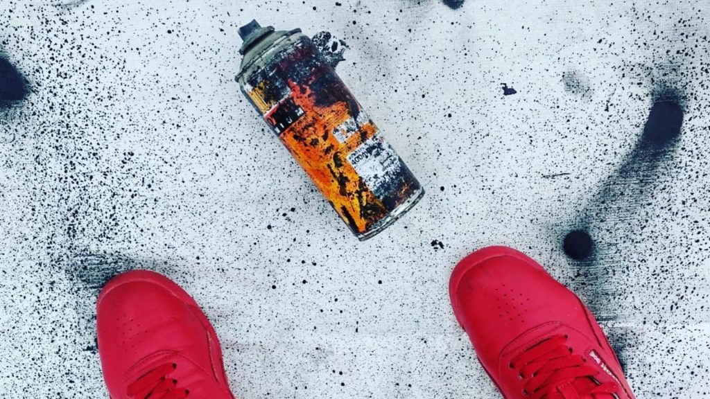 An overhead view of a can of black spray paint and bright red nike dunks. There is spatters of spray paint on the ground