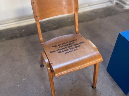 Wooden Chair An adults chair with no arms, and wooden legs, a wooden top backrest and wooden seating base. Black writing saying: "Why are you so shocked that i'm educated? Take a moment and think about what that says about you not me!" This chair is situated in St Johns Church in Peterborough. Its on a concrete floor with a blue information plinth to the right of it.