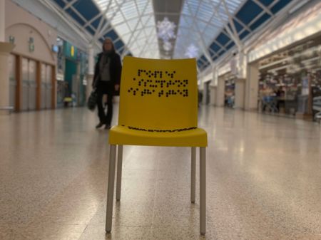 Yellow Adults Chair An adults plastic yellow chair without arms, metal grey legs, a yellow plastic backrest and base. Black brail writing on the backrest of the chair, and writing on the back o fthe backrest saying: People's assumption is that they know more than me because they can see!' The chair is based as Serpentine Green Shopping Centre in Peterborough. The chair is on the smooth stone floor in the shopping centre with shops blurry in the background.