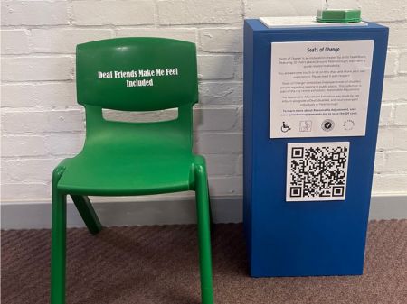 Green chair A small green childrens chair with 4 green legs, a plastic backrest and strong plastic seating base. White writing saying: Deaf friends make me feel included." This chair is situated in the Nova primary school in the hallways with a white painted brickwall behind the chair, and a blue information plinth to the left of it.