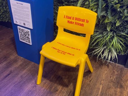 Yellow Chair A small yellow childrens chair with a plastic backrest and strong plastic base. Red writing saying: "I find it difficult to make friends,<br />
I feel like i never quite fit in, I often don't know what to say or do. " This chair is situated in the Dice Box games cafe in Peterborough and stood at the back of the room with the foliage wall behind it and the blue information plinth to the left of it.