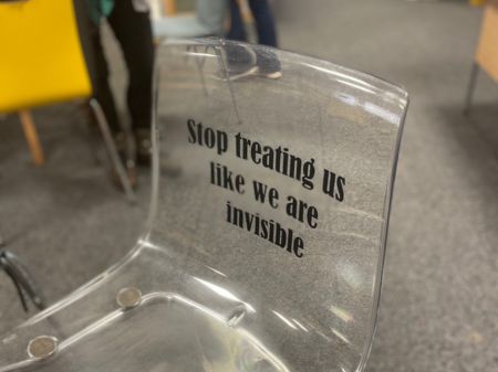 See-through adults chair. An adults plastic clear see-through chair without arms, metal grey legs, a clear plastic backrest and base. Its facing to the left slightly. The writing on the front of the backrest says : "Stop treating us like we are invisible." The chair is on a grey carpet with other chairs in the background around tables. This chair is based at the Dementia Centre in Millfield, Peterborough.