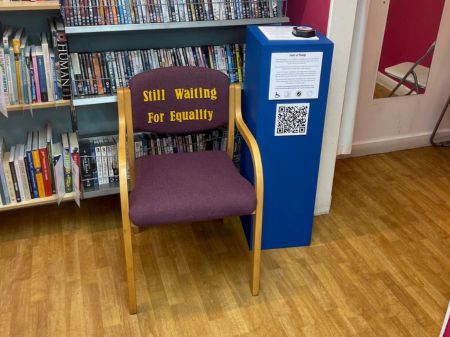 Waiting Room Chair. An adults wooden waiting room chair with arms, wooden legs, a purple cushioned backrest with and a purple cushioned seat. . The writing on the front of the cushioned backrest says : "Still waiting for equality!" The chair is stood on the wooden floor in the Scope Charity Shop. With CD's and DVD's on the wall behind and the plinth to the right.