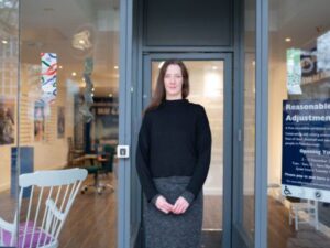 Artist Fae Kilburn standing in the front entrance doorway of the Reasonable Adjustment Exhibition. Fae has long brown hair, a black jumper and a grey skirt. Her hands are clasped in front of her. The front of the building is mainly large glass windows either side of Fae and aglass door behind Fae, although the exhibition is hard to see from the outside.