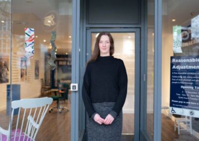 Artist Fae Kilburn standing in the front entrance doorway of the Reasonable Adjustment Exhibition. Fae has long brown hair, a black jumper and a grey skirt. Her hands are clasped in front of her. The front of the building is mainly large glass windows either side of Fae and aglass door behind Fae, although the exhibition is hard to see from the outside.