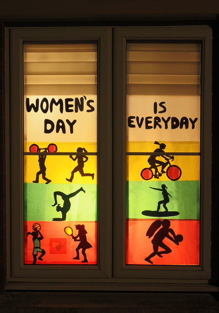 A designed window with Layers of different colours. Red at the bottom, then green above an then yellow above and then white at the top. Silhouette cut outs of women playing different sports accross each colour. "Womens Day if Everyday" written across the top.