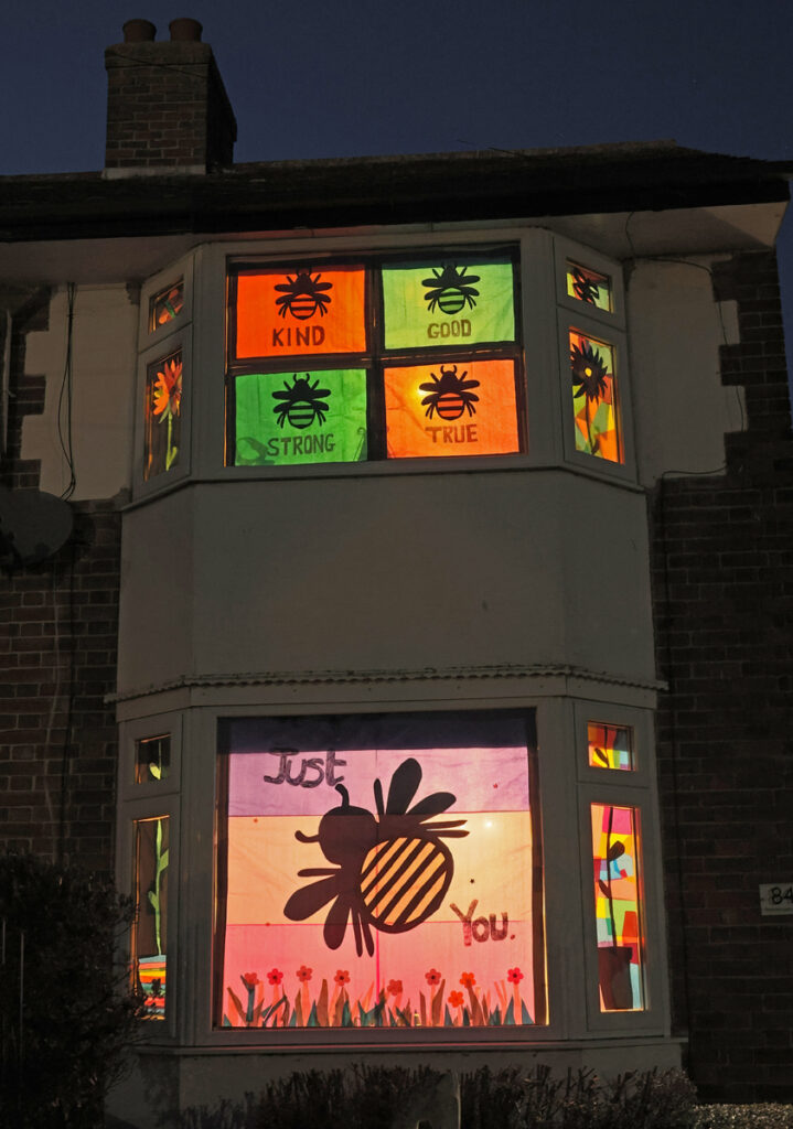 ground floor window decorated with a giant Bee silhouette and a pink, yellow and light purple background. the first floor decorated window has at least 4 smaller bees with values written underneath them and different coloured backgrounds.