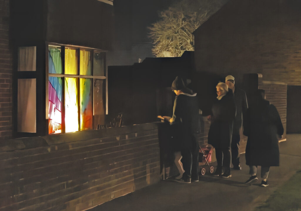 Various people on the right stood at a small wall looking over it at a house on the left with decorated bay window. Its night and the windows are glowing. The window design has a yellow, blue purple and red block coloured windows (Although its not clear what the design is).