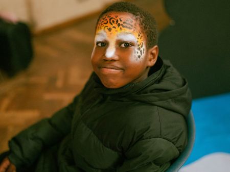A young black child smiles at the camera, showing off his face paint. His forehead, nose, and cheeks have been painted to resemble a cheetah.