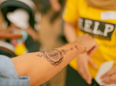 A photo of a person's arm who has just had a henna design drawn onto it. The henna is an intricate mandala pattern on the forearm. The person who has just done the henna is wearing a bright yellow t-shirt which has the word H.E.L.P printed across it in white text with a black background.