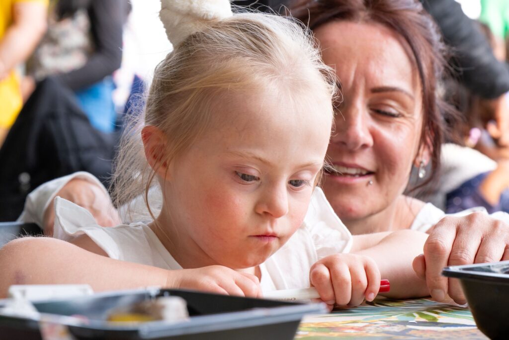 In the Creative dome at the Peterborough Celebrated Festival.... A close up of a fair haired young girl - her hair in a ponytail sat at a table, who is concentrating and looking down whilst making something. Her mum is behind her smiling.