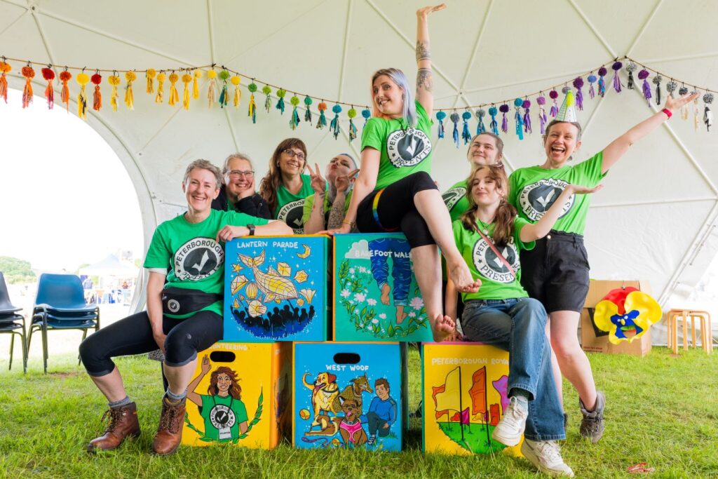 In the Creative dome at the Peterborough Celebrated Festival.... is the end of the first day and the Peterborough Presents team all in their green tshirts are together facing the camera and smiling. Some sat on large wooden cubes and some standing behind them.