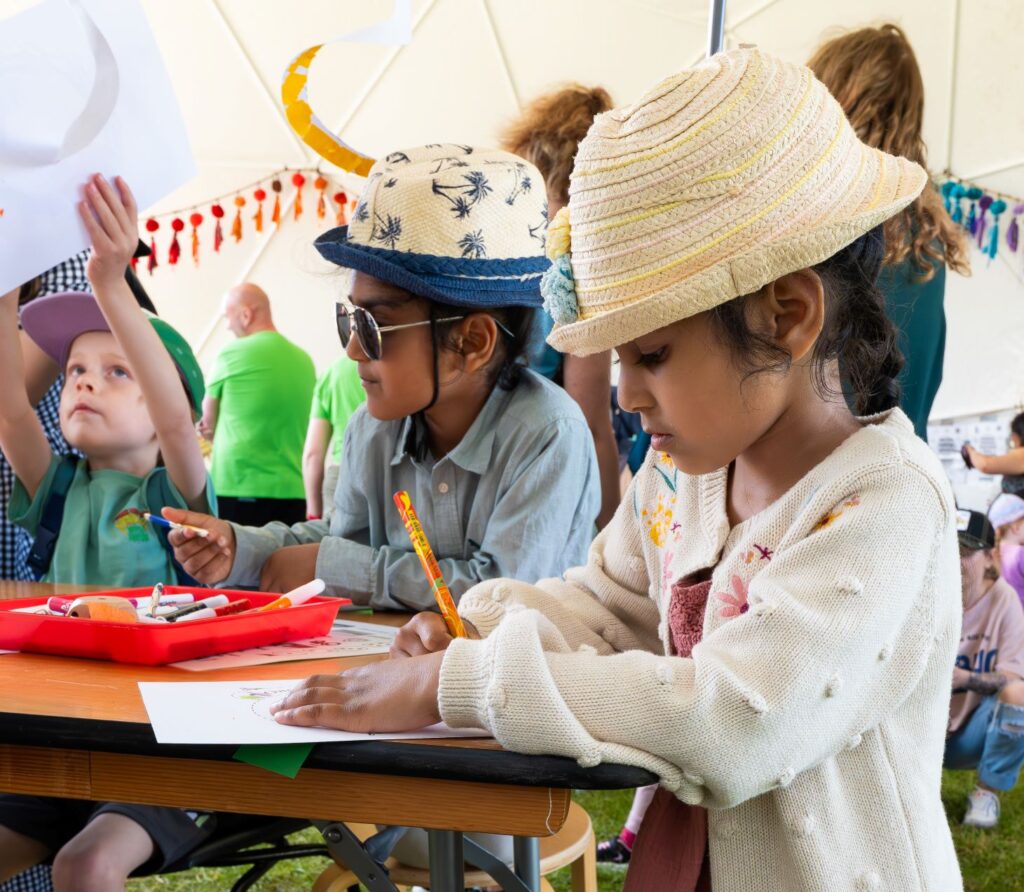 In the Creative dome at the Peterborough Celebrated Festival.... 2 young children and boy and a girl with summer hats on are stood at a table colouring in their story spirals.