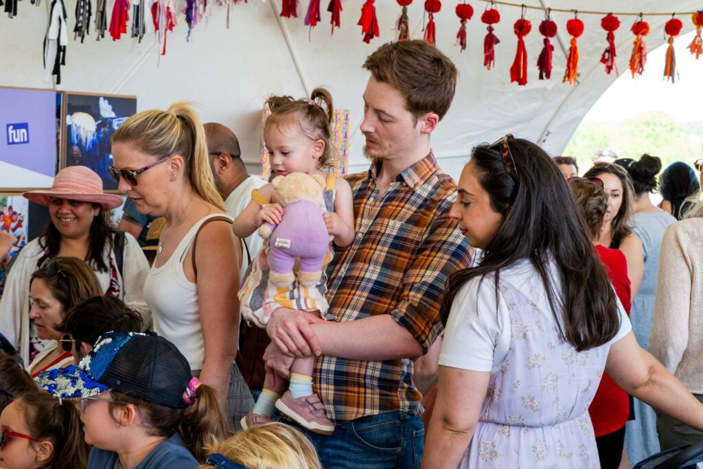 In the Creative dome at the Peterborough Celebrated Festival.... a couple stood together, and the dad is holding her in his arms and looking at her as she holds her teddy and looks at the table. People in the background of the dome.