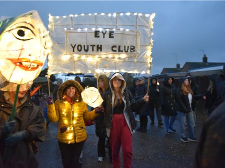 Young people smiling at the camera holding a paper lantern sign between them which reads 'eye youth club'. It is framed in lit up fairylights. There are crowds seen in the background.