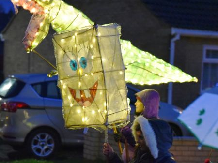 A child carrying a paper lantern in the shape and design of cartoon character Spongebob Squarepants. It's illuminated from within. There is another lantern in the background in the shape of a dinosaur.