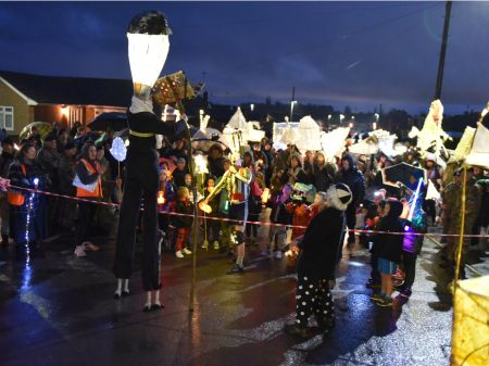 Crowds of people at eye lantern parade stood with their lanterns illuminated looking at a tall figure wearing stilts performing. 