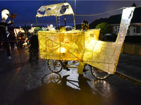 A large hand made paper lantern in the shape of a train, with a bicycle peaking out from beneath. The lantern is well lit from within and has someone within the structure. 