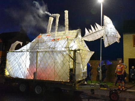 Large paper lantern structure in the shape of the "eye monster". The lantern is white and is lit from within with fairly lights. There is smoke coming from it's chimney on it's back. It's on a trailer. 