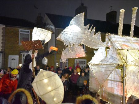 Large paper lantern structure in the shape of the "eye monster". The lantern is white and is lit from within with fairly lights. There is smoke coming from it's chimney on it's back. It's on a trailer.  There are crowds around it on the street. It is being fed a 'red brick' lantern. 