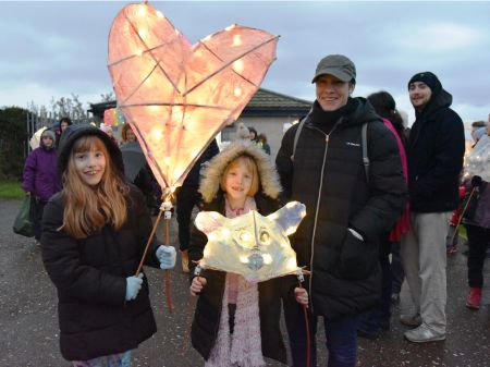 Two young people and an adult smiling whilst the two young people are carrying their homemade paper lanterns in the shape of a heart and a animal face.