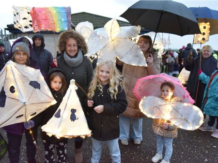 A group of people of varying ages posed with their hand made paper lantern in various shapes and sizes. The people have hoods up and are holding umbrellas and their lanterns are brightly lit from within.