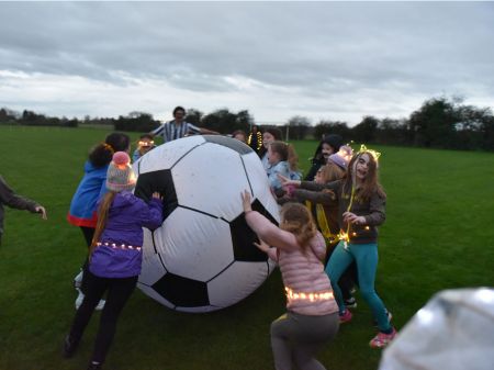 A group of young people, some of whom are wearing fake moustaches and fairylights, playing with a giant inflatable football in a field. Someone dressed as a referee can be seen behind the ball.