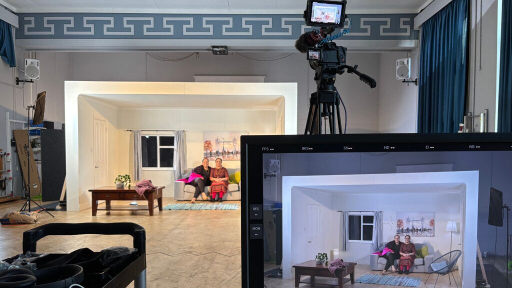 Millfield of Dreams film set. The film monitor and camera is in the right foreground, and in the background are two Indian women sat on the on-set sofa, smiling.