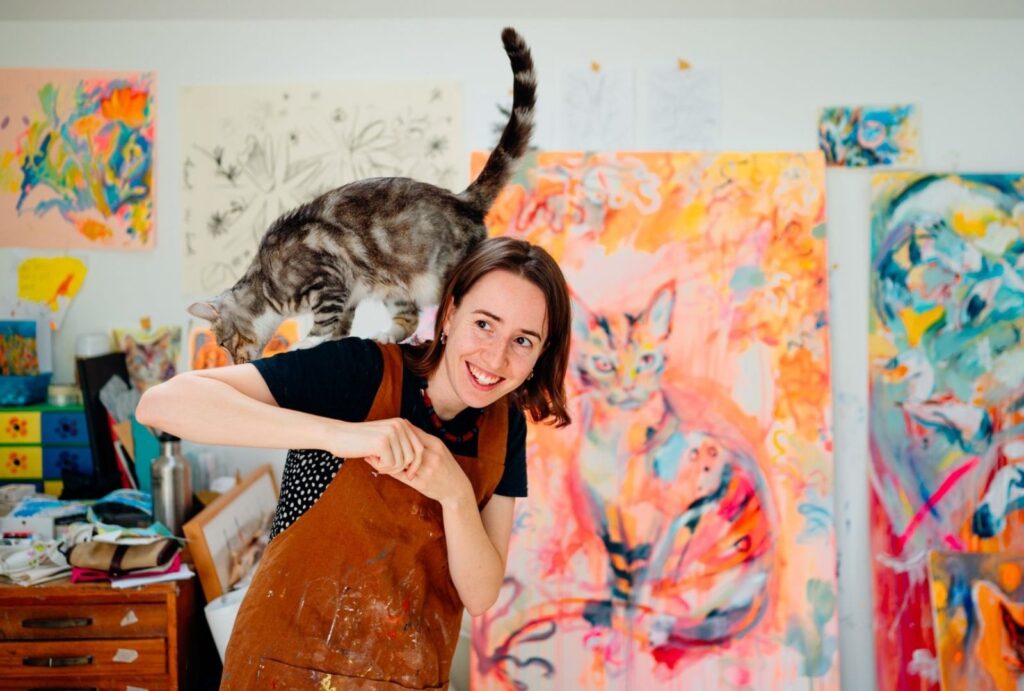 An image of Emily Bowers, a local artist. In the image Emily is smiling whilst a cat playfully climbs on her shoulders. Emily is standing in her painting overalls in what appears to be a colourful painting studio.