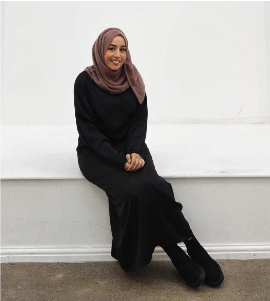An image of local artist Fasiha Ashia sitting on a white side board. She is smiling.